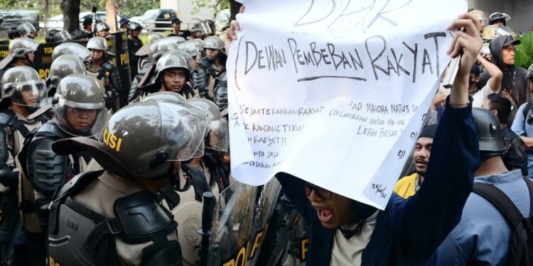 Tiga Orang Masih Hilang Usai Demonstrasi Agustus 2025 Seorang mahasiswa yang membentangkan poster protes terhadap DPR di hadapan polisi dalam aksi unjuk rasa Agustus 2025 di Jakarta. (Foto: Iqro Rinaldi via Unsplash)